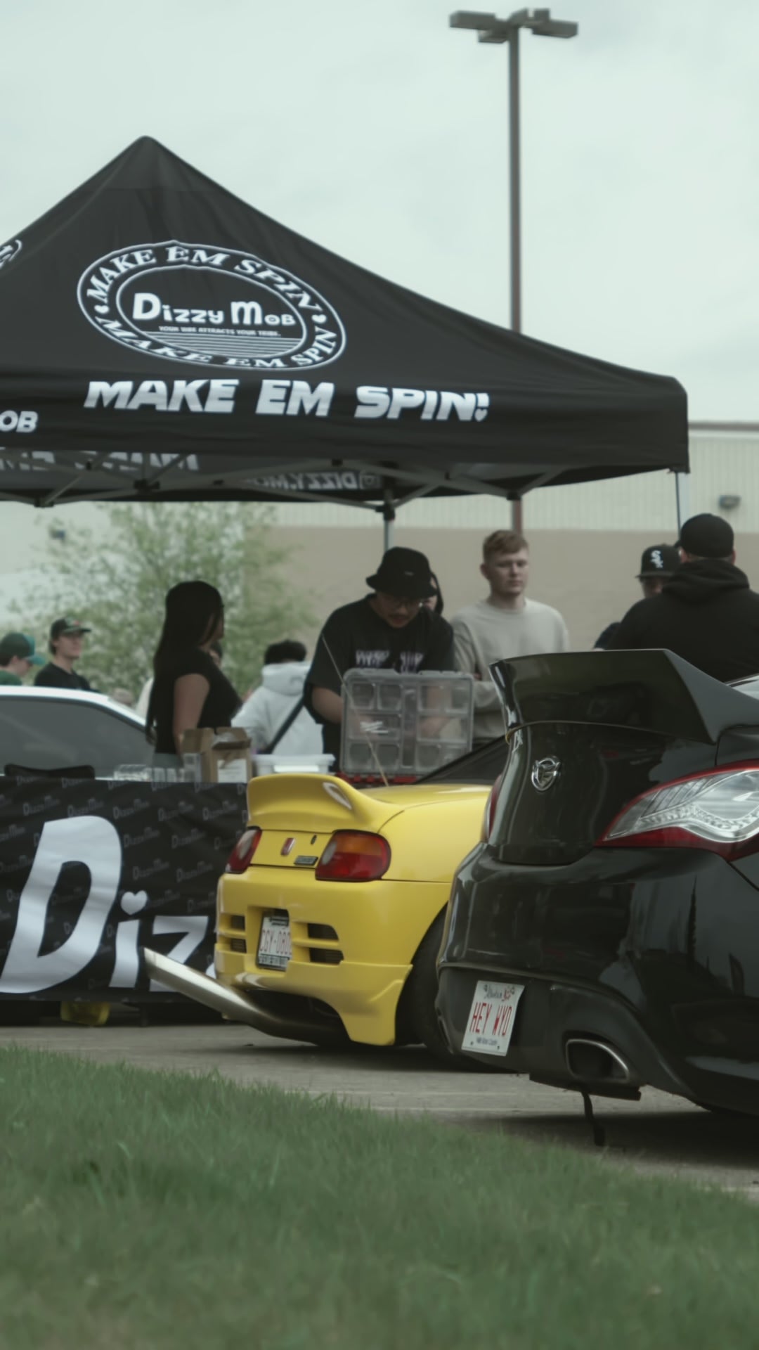 Load video: A video showcasing Dizzy Mob at a local car show in a parking lot filled with modified cars, vendors and spectators walking around looking at cars, talking to friends and shopping at the booth